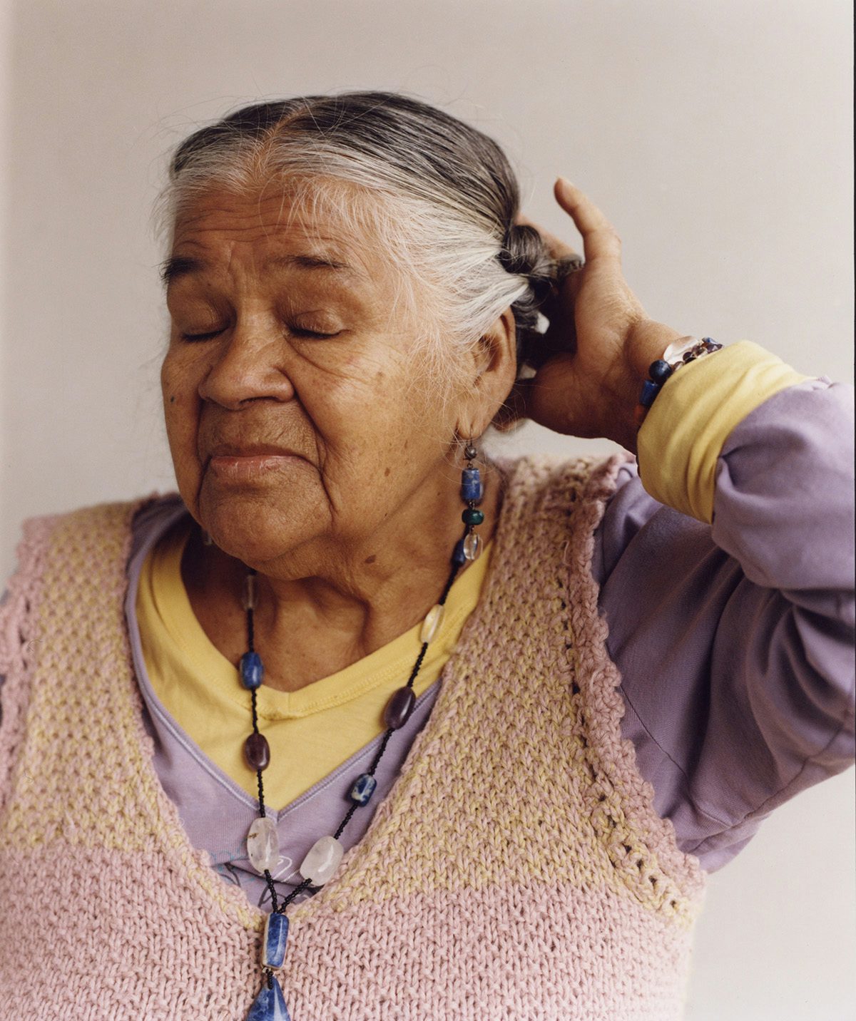 Portrait photo by Ana Margarita Flores of a person with their eyes closed and their hand held up towards their grey braided hair, and wearing layers of clothes in yellow and pale purple