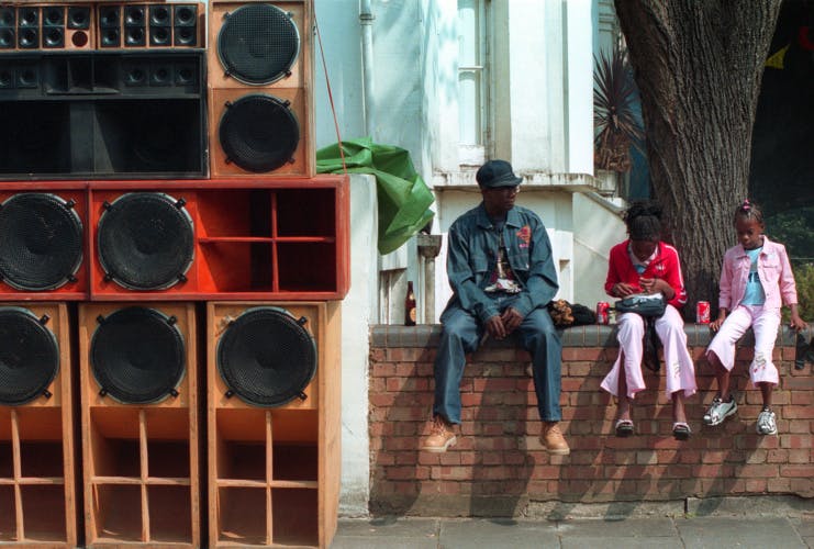 An adult and two young people sat on a wall next to a speaker stack in the street