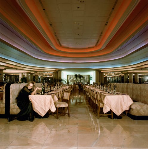 Photograph of the model Twiggy sat on a booth in at the front of a row of tables in a large restaurant space with a ceiling covered in multicoloured curving patterns