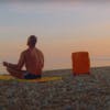 A muscular topless man wearing swim shorts sat on a towel on a pebble beach at sunset, with an orange Floyd suitcase next to him