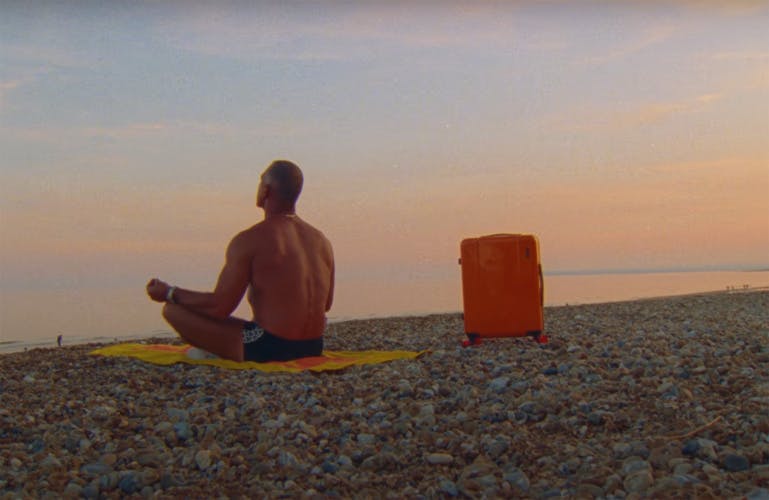A muscular topless man wearing swim shorts sat on a towel on a pebble beach at sunset, with an orange Floyd suitcase next to him