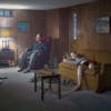 A person sat in an armchair and another person lounging on a mustard sofa in a wood panelled room, both gazing ahead into the distance