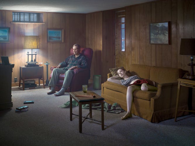 A person sat in an armchair and another person lounging on a mustard sofa in a wood panelled room, both gazing ahead into the distance