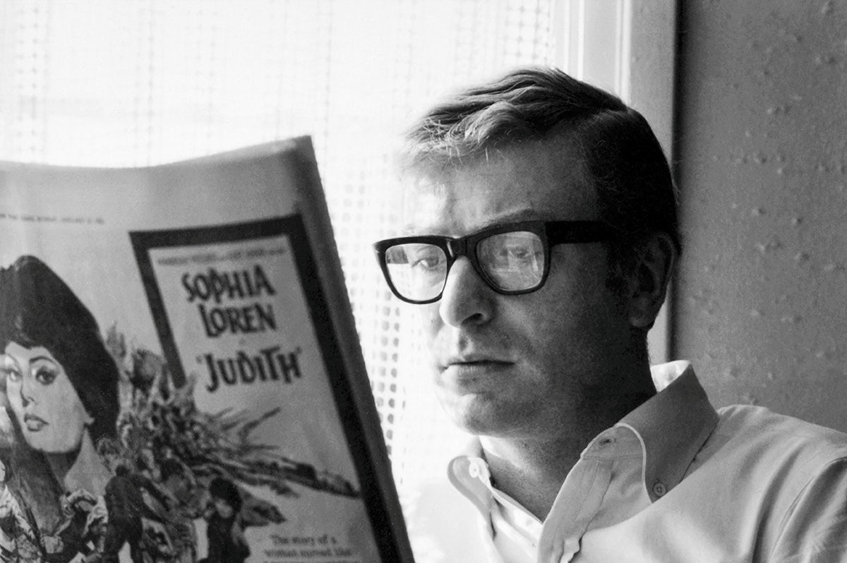 Black and white photo of Michael Caine wearing thick rimmed glasses and a shirt reading a publication with a Sophia Loren advert visible on the back page