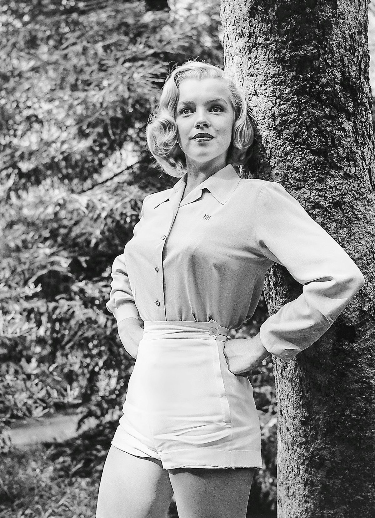 Black and white photo of Marilyn Monroe wearing shorts and a shirt with her hands placed on her hips standing next to a tree