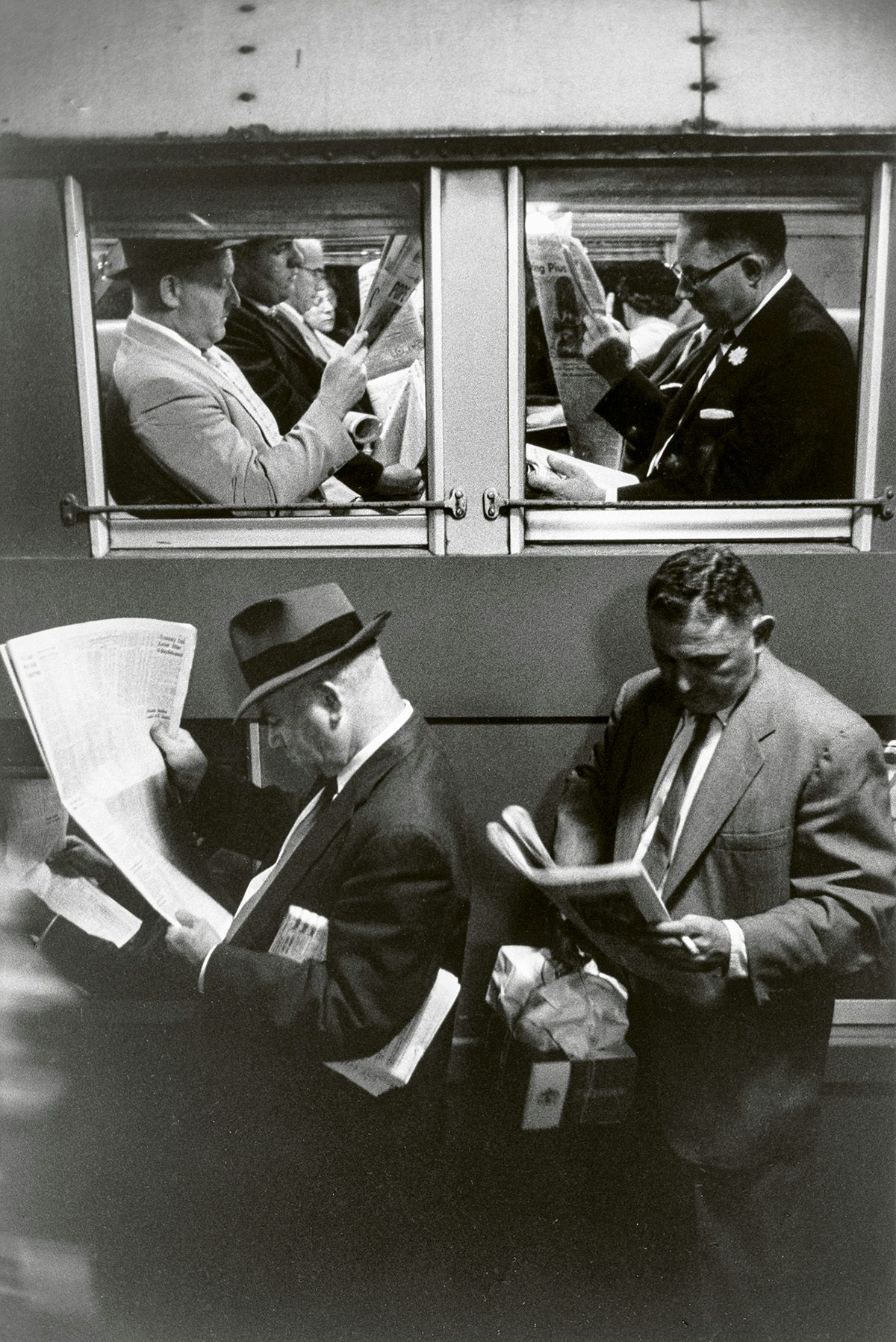 Black and white photo by Louis Stettner of suited men reading newspapers on a train platform, and more reading newspapers sat on the train behind them