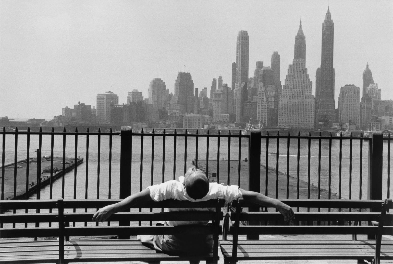 Black and white photo by Louis Stettner of a person leaning back on a bench with the New York skyline in the distance