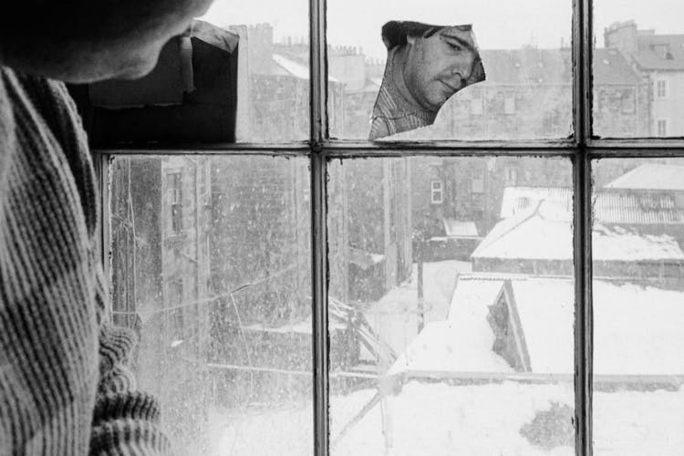 Black and white photo of a person peering through a dusty window, their reflection cast in a cracked piece of mirror resting on one of the panes