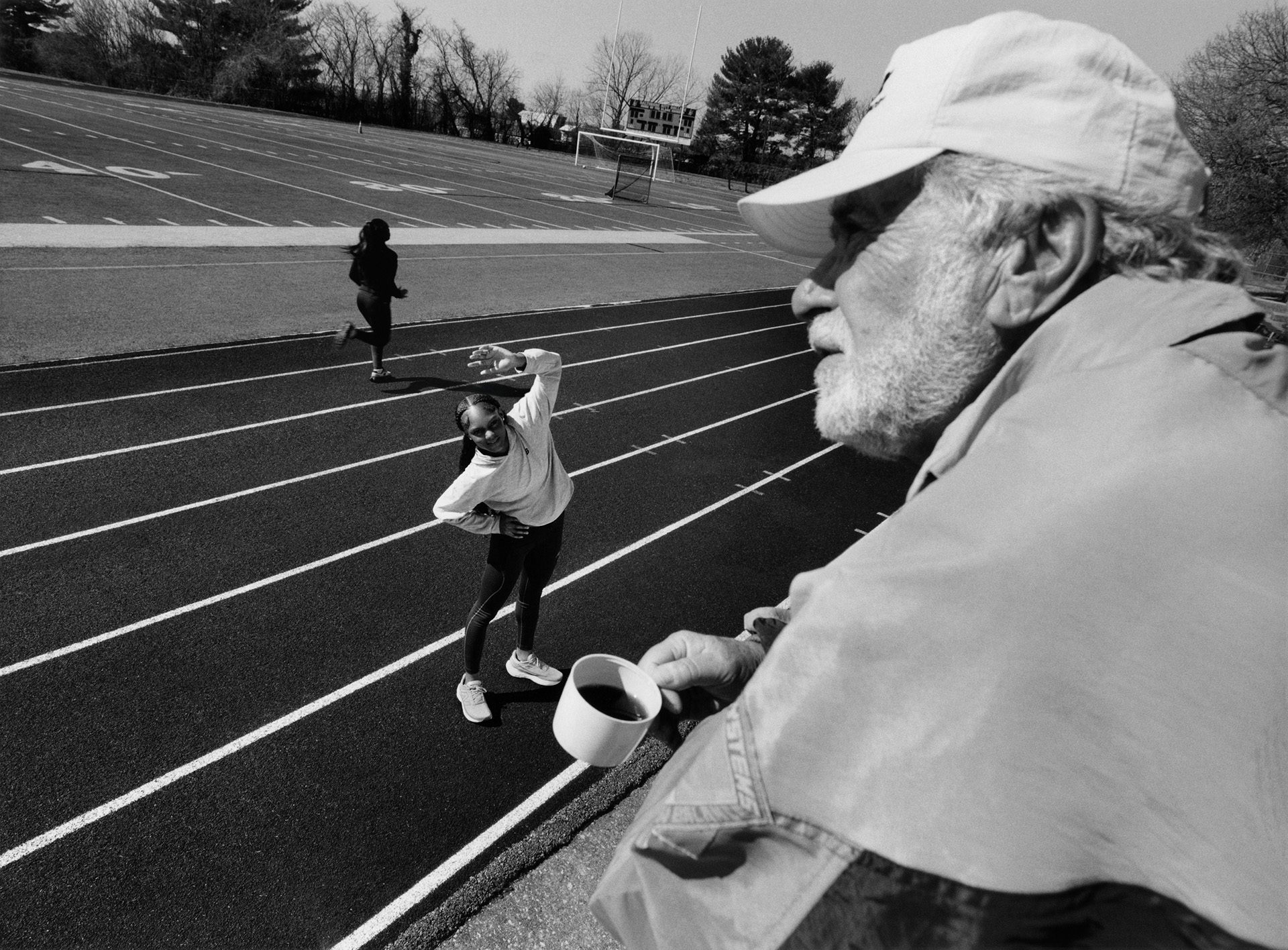Black and white shot of an older person wearing a cap and track sports jacket holding a mug, looking out over an outdoor running track where a younger person is stretching