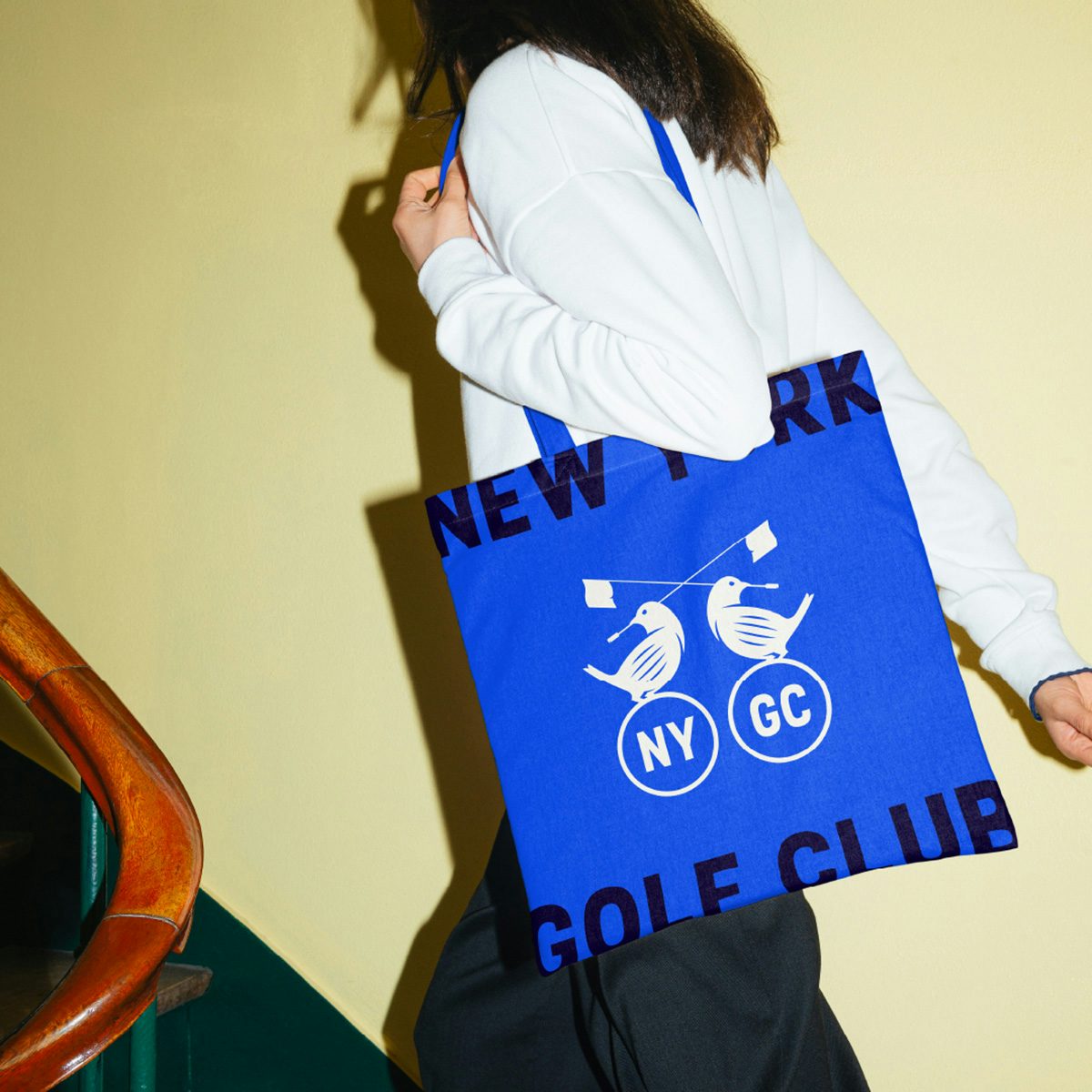 A person wearing a tote bag featuring the New York Golf Club bird emblems holding a golf flag in their beaks, and the initials NY GC