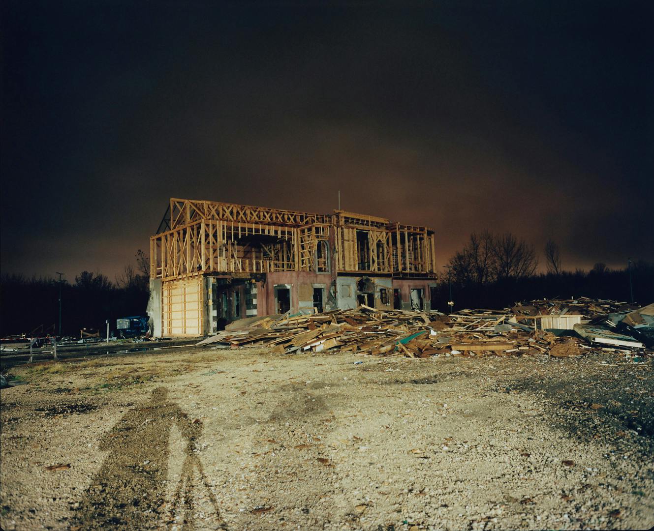 Exterior of a film set in Budapest with the skeleton of the building visible from the outside, photographed at night. The shadow of photographer Yorgos Lanthimos and his tripod is visible on the ground in front of the building